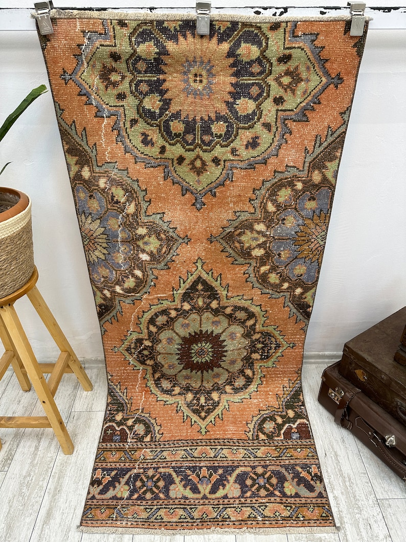 May include: A vintage Turkish runner rug with a faded peach background and intricate floral patterns in shades of green, brown, and blue. The rug has a fringed edge and a geometric border.