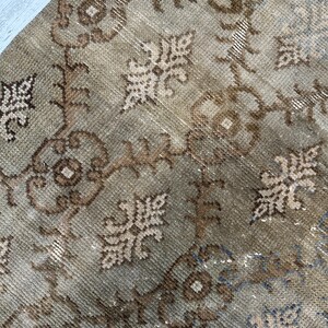 May include: A woven rug featuring a repeating pattern of brown and beige floral motifs and geometric shapes. The textile displays a distressed, weathered texture with visible knots and a color palette consisting of muted earth tones and soft blue.