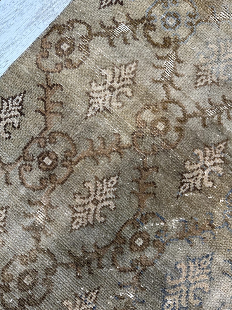 May include: A woven rug featuring a repeating pattern of brown and beige floral motifs and geometric shapes. The textile displays a distressed, weathered texture with visible knots and a color palette consisting of muted earth tones and soft blue.