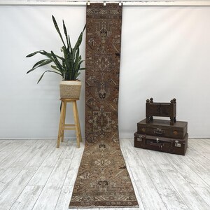 May include: A long, narrow runner rug featuring a complex geometric pattern in shades of brown, tan, and cream. The rug displays intricate floral and tribal motifs throughout its length, with a symmetrical design and a fringed edge at the bottom.
