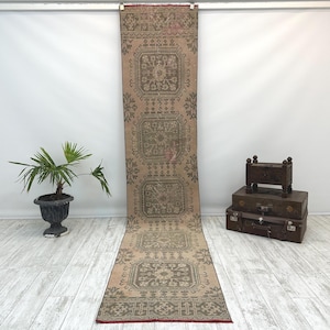 May include: A long, narrow runner rug featuring a muted tan background with repeating dark gray geometric and floral motifs arranged in square panels. The rug has a thin red border along the bottom edge and a textured, woven appearance.