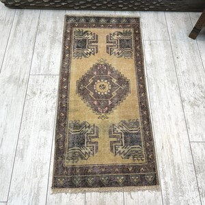 May include: A rectangular, vintage-style rug with a central diamond pattern in shades of brown, tan, and pink. The rug features geometric designs at the top and bottom, framed by a dark brown border. The rug is approximately 24 inches long.