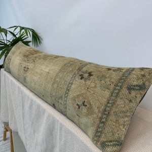 May include: A long, rectangular lumbar cushion featuring a weathered, textured surface in muted shades of beige, tan, and faded sage green. The fabric displays a repeating geometric pattern with worn, distressed details across the entire length.