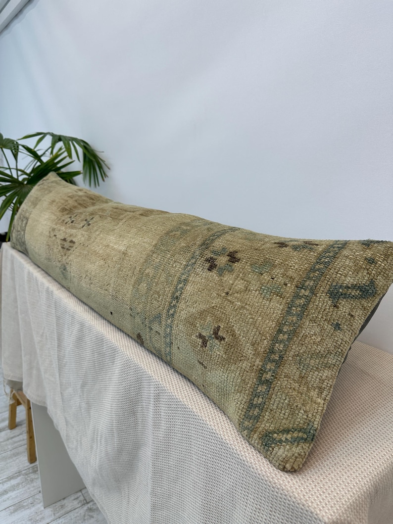 May include: A long, rectangular lumbar cushion featuring a weathered, textured surface in muted shades of beige, tan, and faded sage green. The fabric displays a repeating geometric pattern with worn, distressed details across the entire length.