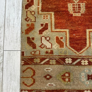May include: A vintage rug with a rust-colored background and a geometric pattern in shades of brown, beige, and white. The rug has a fringe border.