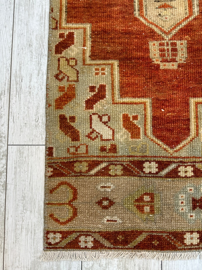May include: A vintage rug with a rust-colored background and a geometric pattern in shades of brown, beige, and white. The rug has a fringe border.