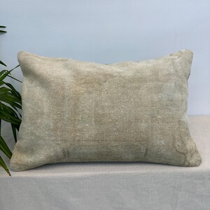 May include: A rectangular throw pillow featuring a weathered, light beige woven fabric with a subtle, faded texture. The surface displays a neutral, earthy tone with slight variations in colour density across the front, creating a worn appearance.