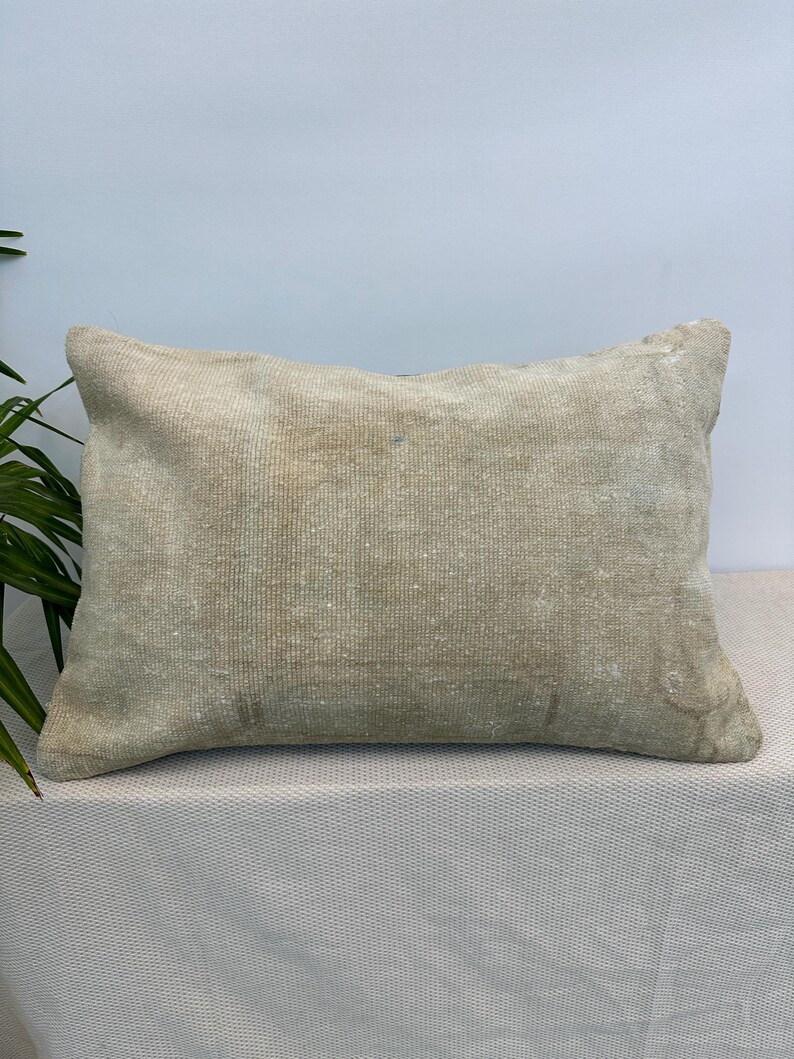 May include: A rectangular throw pillow featuring a weathered, light beige woven fabric with a subtle, faded texture. The surface displays a neutral, earthy tone with slight variations in colour density across the front, creating a worn appearance.