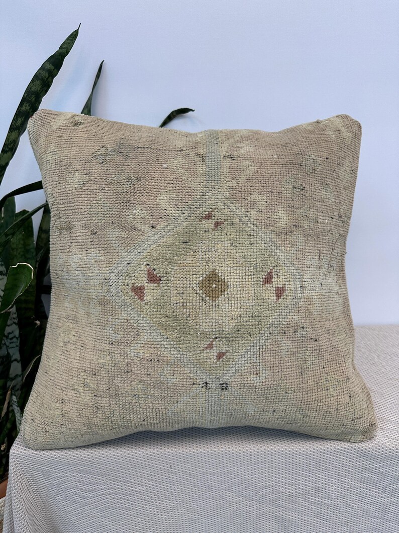 May include: A square throw pillow featuring a woven, textured fabric in muted beige and light tan tones. The center displays a diamond-shaped geometric pattern in faded green, accented by small, scattered red and brown details throughout.