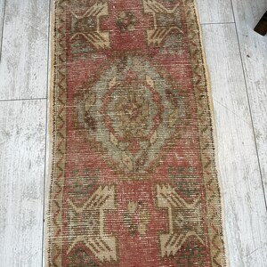 Puede incluir: Una alfombra rectangular de estilo vintage con una paleta de colores rojo y beige descoloridos. La alfombra presenta patrones geom&eacute;tricos y un dise&ntilde;o de medall&oacute;n central. Los bordes est&aacute;n acabados con flecos. La alfombra est&aacute; colocada sobre un suelo de madera clara.