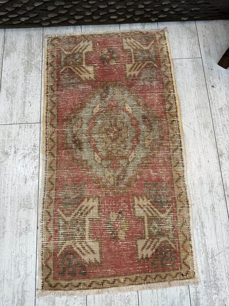 Puede incluir: Una alfombra rectangular de estilo vintage con una paleta de colores rojo y beige descoloridos. La alfombra presenta patrones geom&eacute;tricos y un dise&ntilde;o de medall&oacute;n central. Los bordes est&aacute;n acabados con flecos. La alfombra est&aacute; colocada sobre un suelo de madera clara.
