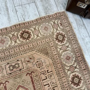 May include: A beige rug with a brown and pink geometric pattern. The rug has a fringe border and is made of a soft, woven material.