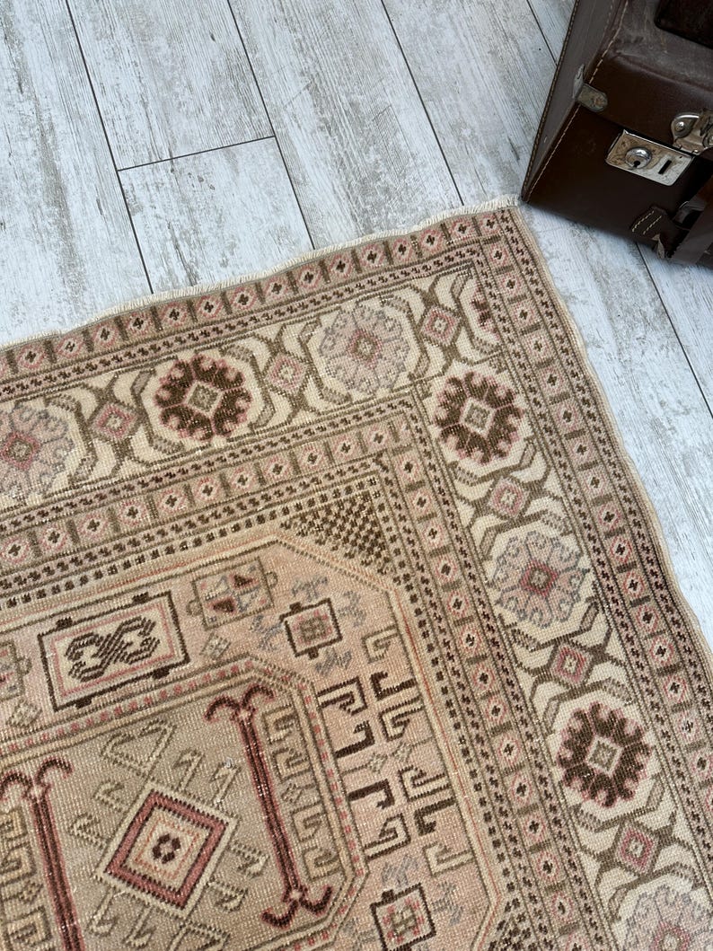 May include: A beige rug with a brown and pink geometric pattern. The rug has a fringe border and is made of a soft, woven material.