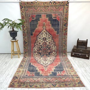 May include: A large, rectangular rug with a central medallion design. The rug features a red field with navy blue and beige accents. The border is a mix of green, beige, and red. The rug is hanging against a white wall.