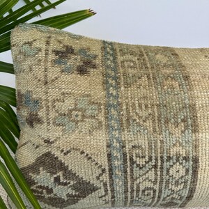 May include: A rectangular throw cushion featuring a textured, woven surface with a muted colour palette of beige, soft blue, and dark brown. The design displays intricate geometric patterns and floral motifs arranged in vertical panels across the fabric.