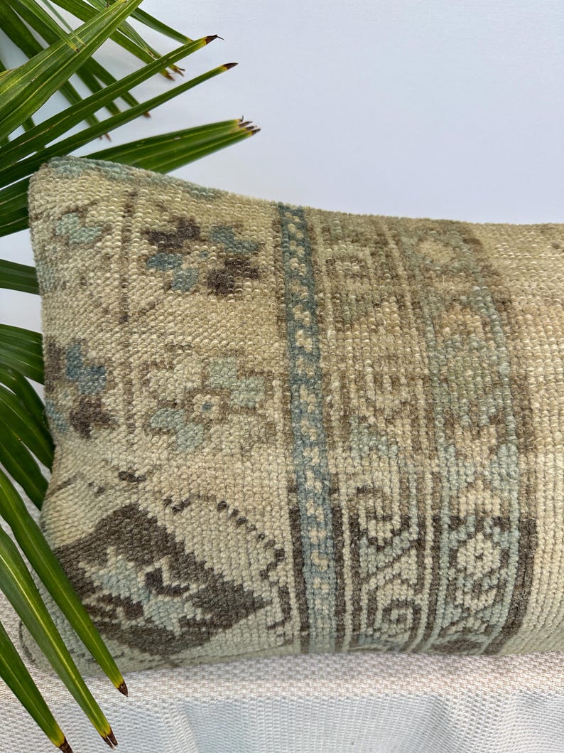May include: A rectangular throw cushion featuring a textured, woven surface with a muted colour palette of beige, soft blue, and dark brown. The design displays intricate geometric patterns and floral motifs arranged in vertical panels across the fabric.