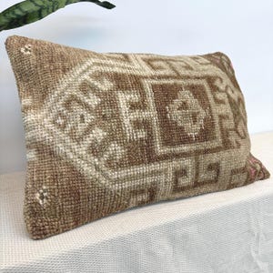 Puede incluir: Un cojín rectangular con una superficie tejida y texturizada en tonos marrón claro, beige y crema. El cojín presenta un patrón geométrico prominente con motivos centrales de diamantes y ángulos, con una construcción de nudos densos.