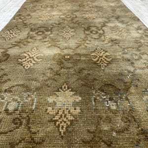 May include: A rectangular runner rug featuring a muted olive green and brown color palette. The surface displays a repeating geometric pattern with stylized floral motifs in lighter beige tones, finished with a short fringe along the edge.