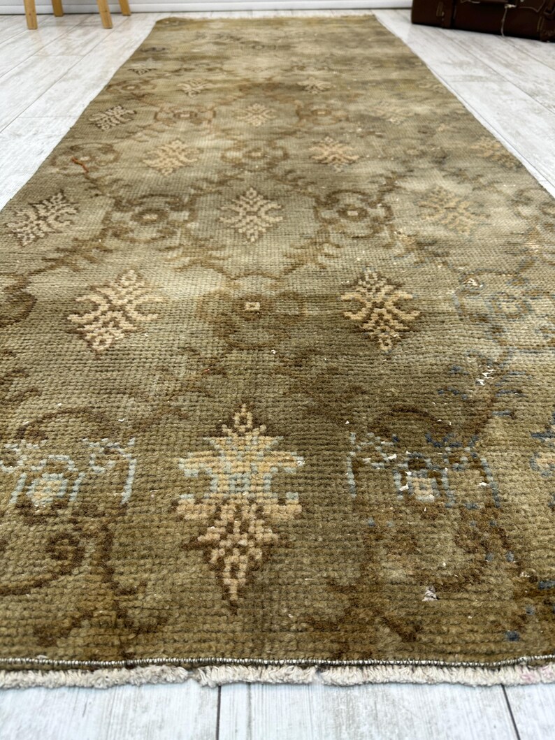 May include: A rectangular runner rug featuring a muted olive green and brown color palette. The surface displays a repeating geometric pattern with stylized floral motifs in lighter beige tones, finished with a short fringe along the edge.