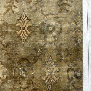May include: A woven rug featuring a muted olive green base with a repeating pattern of dark brown and light tan floral motifs. The design includes intricate symmetrical shapes and scrolling vine elements distributed across the textured surface.