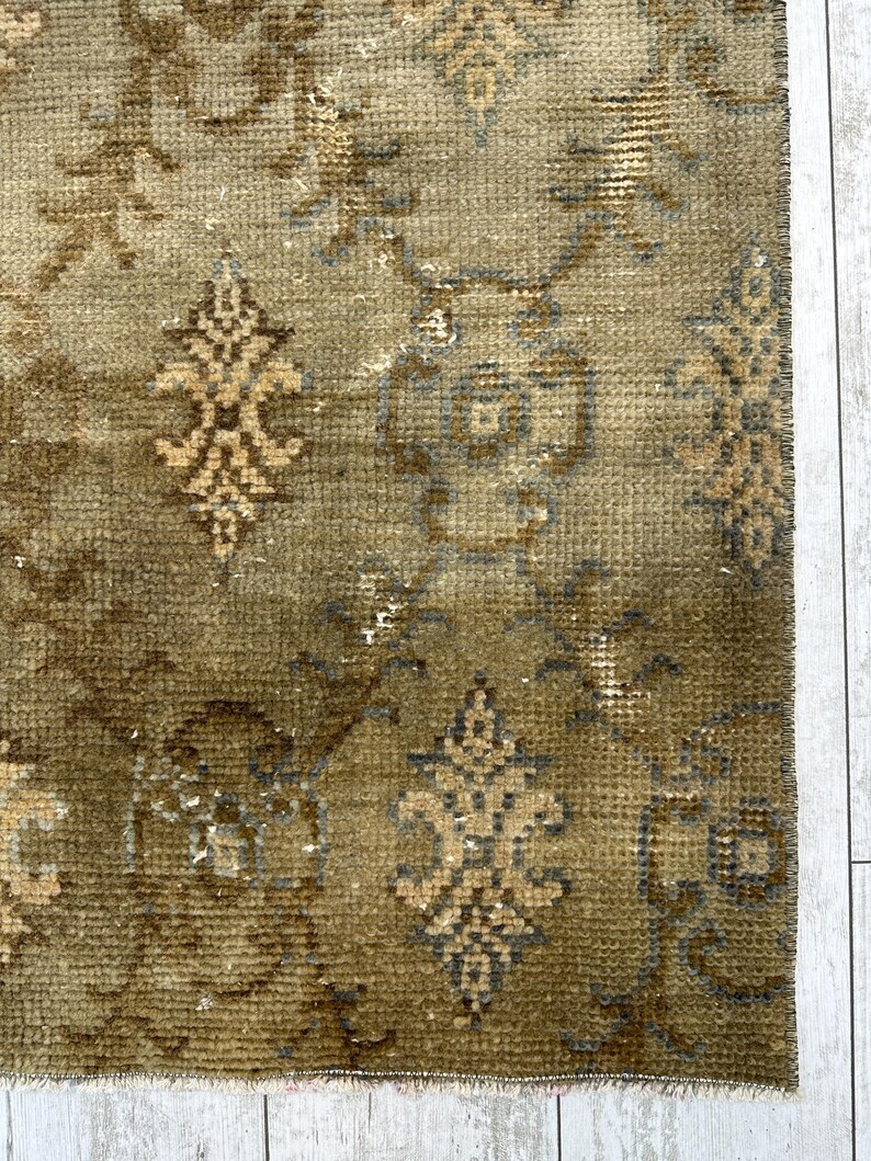 May include: A woven rug featuring a muted olive green base with a repeating pattern of dark brown and light tan floral motifs. The design includes intricate symmetrical shapes and scrolling vine elements distributed across the textured surface.