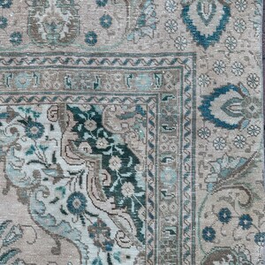 Puede incluir: Una alfombra vintage con fondo beige y intrincados patrones florales en tonos azules y verdes. La alfombra tiene un aspecto descolorido y desgastado, lo que aumenta su encanto antiguo.