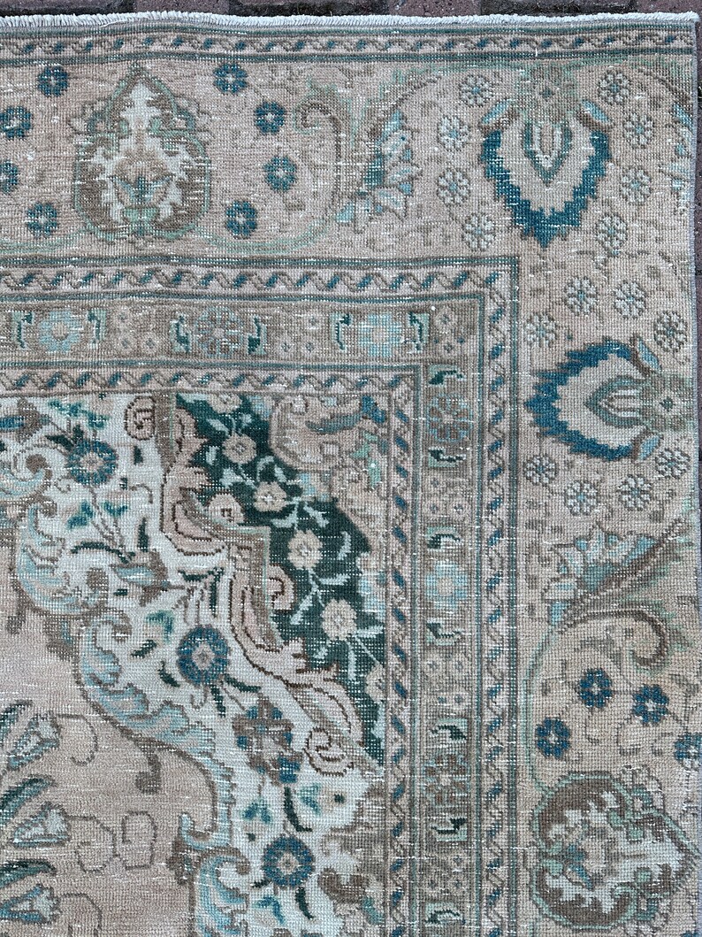 Puede incluir: Una alfombra vintage con fondo beige y intrincados patrones florales en tonos azules y verdes. La alfombra tiene un aspecto descolorido y desgastado, lo que aumenta su encanto antiguo.