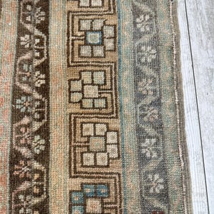 May include: Close-up of a section of a rug with a geometric pattern. The rug features a mix of colors, including brown, beige, blue, and gray. The design includes floral and geometric shapes, creating a textured and detailed appearance. The rug has a fringed edge.