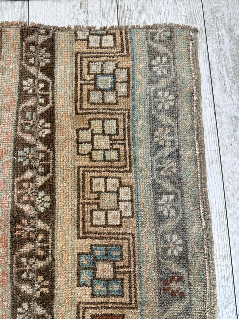 May include: Close-up of a section of a rug with a geometric pattern. The rug features a mix of colors, including brown, beige, blue, and gray. The design includes floral and geometric shapes, creating a textured and detailed appearance. The rug has a fringed edge.