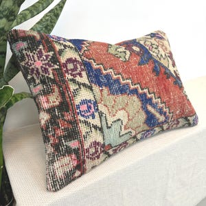 May include: A rectangular throw pillow featuring a woven textile pattern with a distressed appearance. The design includes geometric shapes and floral motifs in shades of rust orange, deep blue, cream, black, and accents of pink and light green.