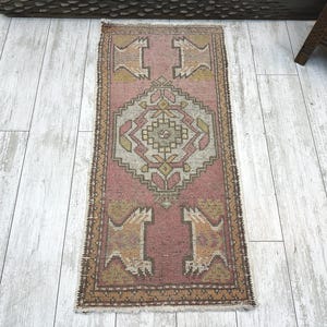 Op de afbeelding: Een rechthoekig vloerkleed met een oudroze achtergrond en een centraal geometrisch medaillon in tinten crème, olijfgroen en beige. Het kleed heeft een decoratieve rand en gestileerde hoekmotieven, afgewerkt met korte franjes aan beide uiteinden.