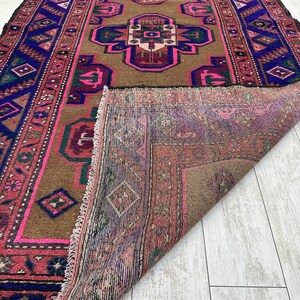 May include: A vintage rug with a brown background and a central design featuring pink, blue, and green geometric shapes. The rug has a fringed edge and is folded in half, revealing the back side.