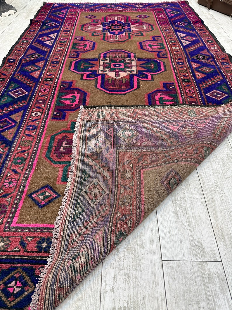 May include: A vintage rug with a brown background and a central design featuring pink, blue, and green geometric shapes. The rug has a fringed edge and is folded in half, revealing the back side.