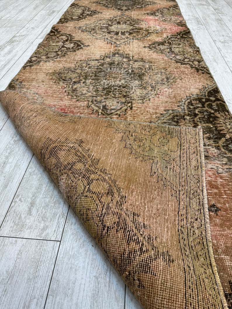 Puede incluir: Una alfombra vintage con un patr&oacute;n floral descolorido en tonos de rosa, marr&oacute;n y verde. La alfombra est&aacute; doblada por la mitad, revelando el reverso, que es de color marr&oacute;n liso.