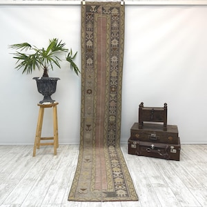 Op de afbeelding: Een lang, rechthoekig tapijt met een centrale roze streep en patroonranden in tinten bruin, beige en groen. Het tapijt wordt verticaal tegen een witte muur getoond. Het ontwerp van het tapijt bevat geometrische en bloemmotieven.