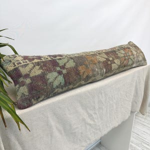 May include: A long, rectangular lumbar pillow featuring a woven textile cover with a faded, multi-colored geometric pattern in shades of deep purple, muted green, and burnt orange. The fabric displays a distressed, weathered texture throughout.