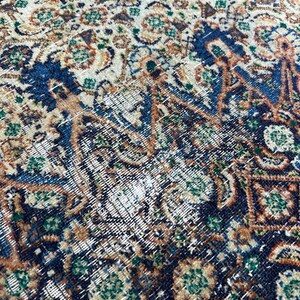 May include: Close-up of a vintage rug with a detailed, intricate pattern in shades of blue, green, brown, and cream. The rug has a worn and faded appearance, with some areas showing signs of wear and tear.