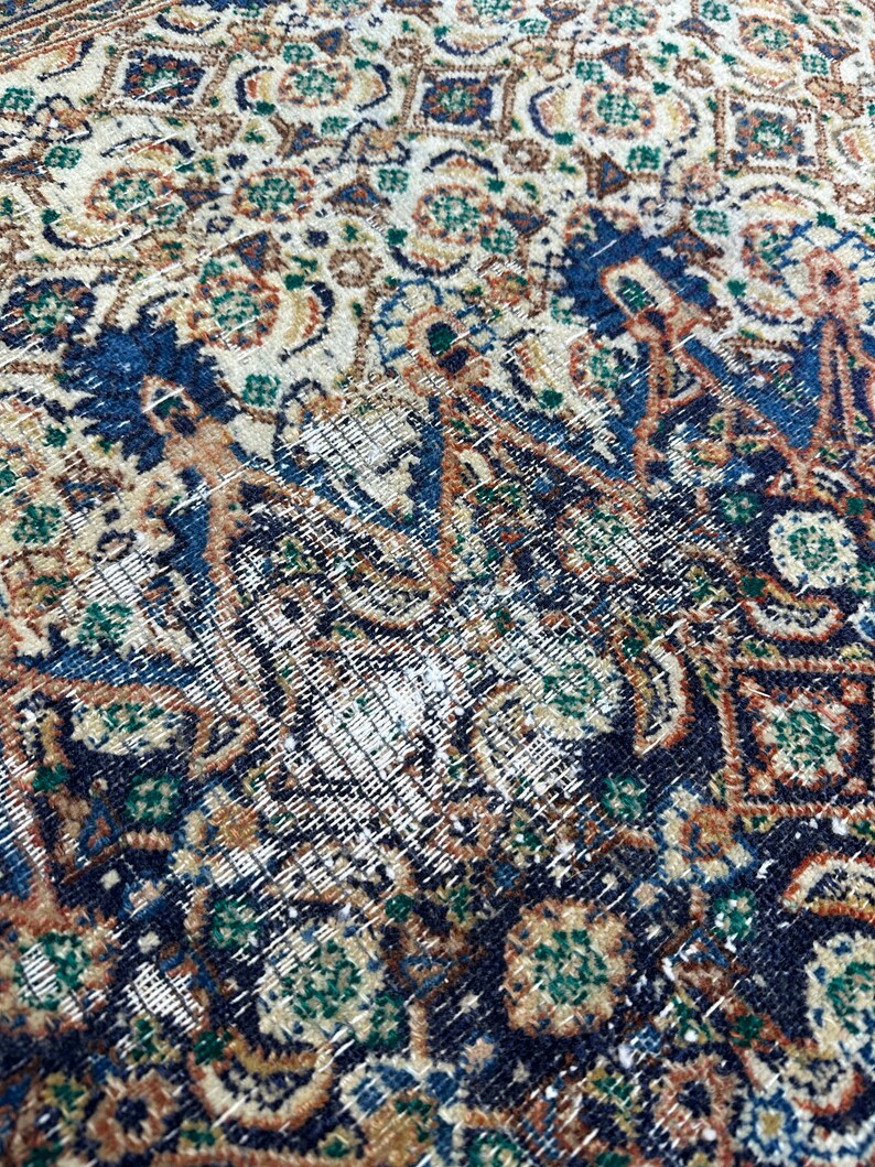 May include: Close-up of a vintage rug with a detailed, intricate pattern in shades of blue, green, brown, and cream. The rug has a worn and faded appearance, with some areas showing signs of wear and tear.