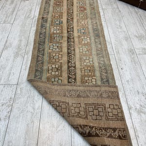 May include: A rectangular, vintage-style rug with a distressed appearance. The rug features a central design of vertical stripes in beige, brown, and muted blue-green tones. The border and ends showcase geometric patterns. The rug is partially rolled up, revealing the underside.