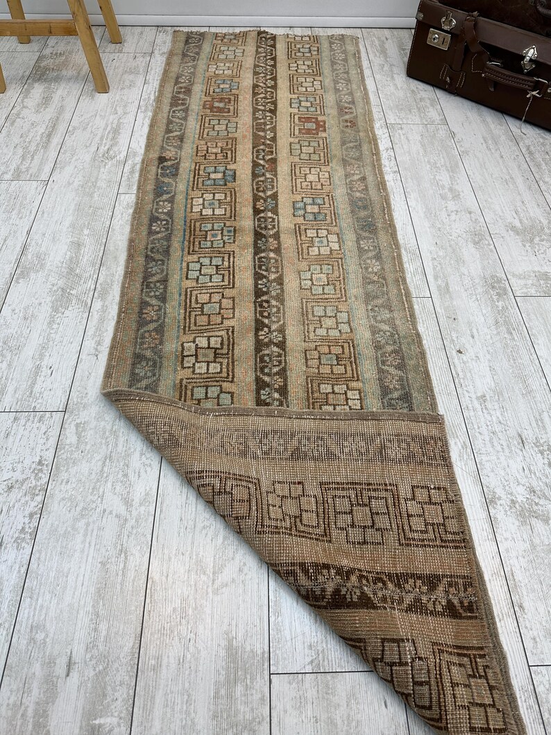 May include: A rectangular, vintage-style rug with a distressed appearance. The rug features a central design of vertical stripes in beige, brown, and muted blue-green tones. The border and ends showcase geometric patterns. The rug is partially rolled up, revealing the underside.