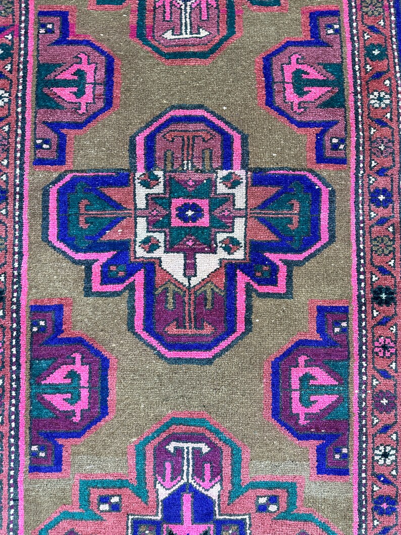 May include: A vintage rug with a brown background and a central design featuring a cross shape with pink, blue, and green accents. The rug has a border with a floral pattern.