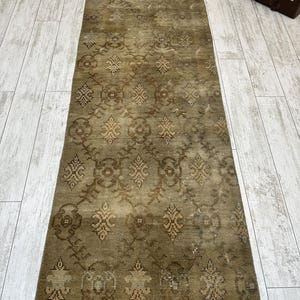 May include: A long, rectangular runner rug featuring a muted olive green and brown color palette. The surface displays a repeating geometric pattern of stylized floral motifs and interconnected diamond shapes, finished with short fringe at both ends.