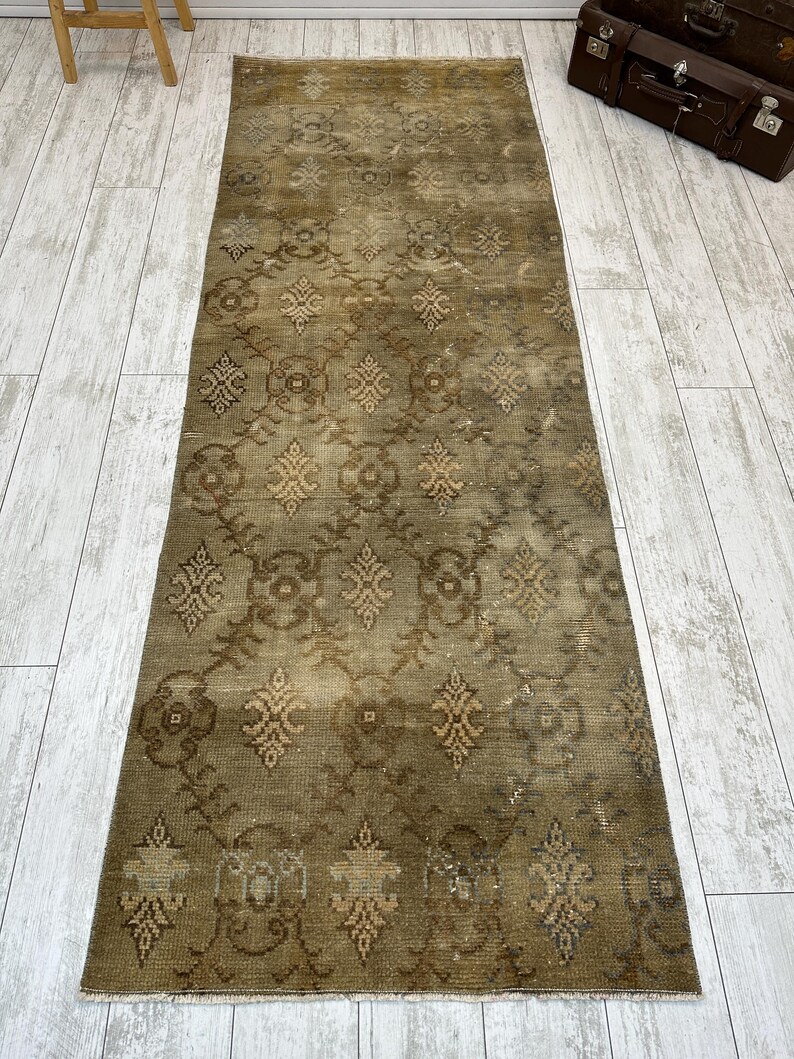 May include: A long, rectangular runner rug featuring a muted olive green and brown color palette. The surface displays a repeating geometric pattern of stylized floral motifs and interconnected diamond shapes, finished with short fringe at both ends.