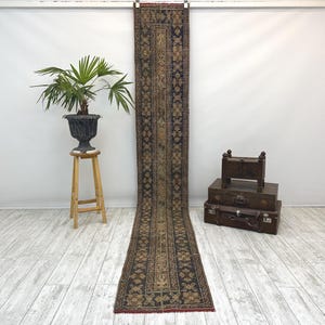 May include: A long, narrow, vintage rug with a dark blue and brown geometric pattern. The rug is displayed vertically, showcasing its intricate design and rich colors. The rug is likely made of wool or a similar material.