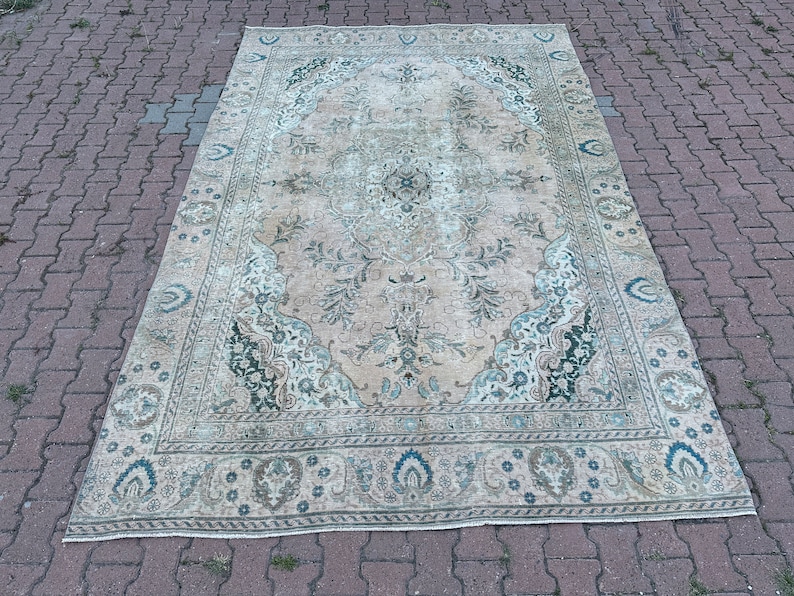 Puede incluir: Una alfombra vintage con fondo crema y un patr&oacute;n floral en tonos verdes y azules. La alfombra tiene un aspecto descolorido y una textura desgastada.