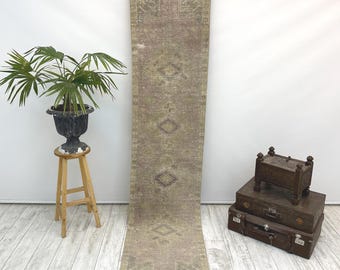 Puede incluir: Una alfombra de pasillo larga y estrecha con una paleta de colores desvaídos y apagados de rosa empolvado, verde salvia y beige. La alfombra muestra una serie de motivos geométricos de diamantes repetidos en el centro, enmarcados por un borde.