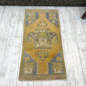 May include: A rectangular area rug featuring a central diamond motif in shades of mustard yellow, slate blue, and muted green. The rug displays geometric patterns and stylized floral elements throughout, finished with a short fringe on both ends.