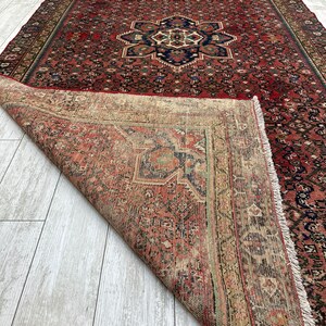 May include: A vintage Persian rug with a red background and intricate floral patterns. The rug is folded in half, revealing the back side, which is a lighter beige color with a similar pattern.