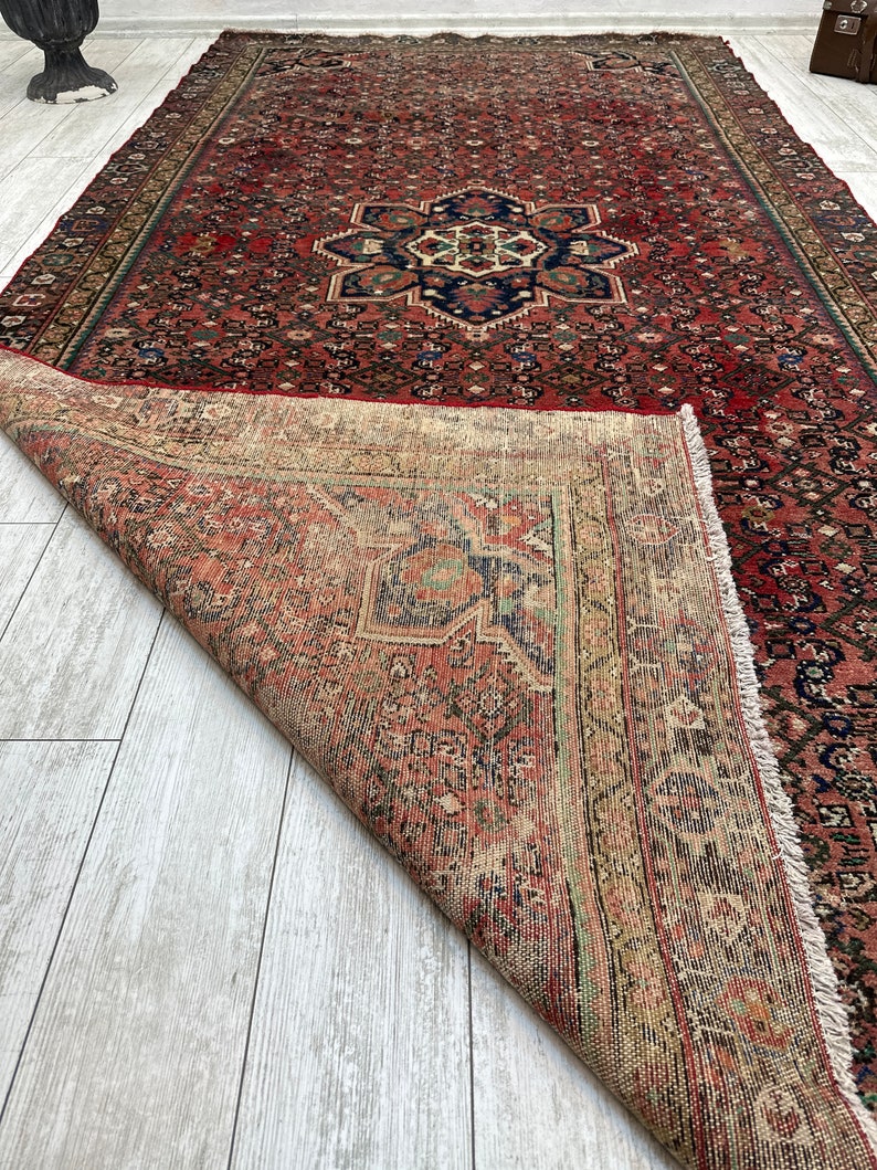 May include: A vintage Persian rug with a red background and intricate floral patterns. The rug is folded in half, revealing the back side, which is a lighter beige color with a similar pattern.