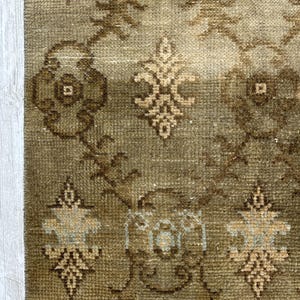 May include: A woven rug featuring a muted olive green and brown color palette with a repeating geometric pattern of stylized floral motifs and interconnected diamond shapes. The texture appears dense and low-pile with a fringed edge at the bottom.
